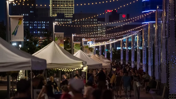 In the night, white tents are set up below light yellow lightbulbs on strings in Scissortail Park.