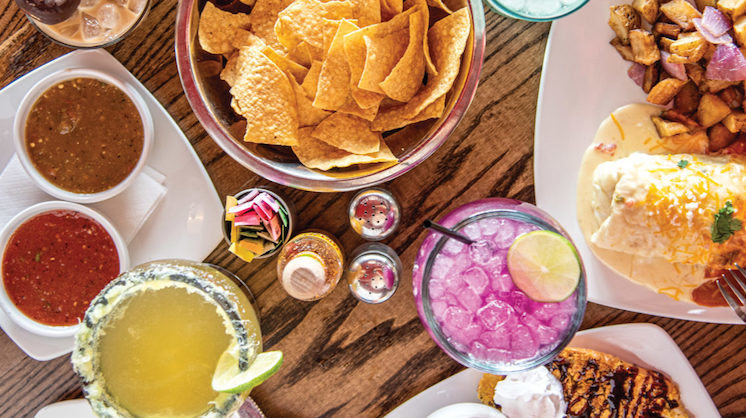 An assortment of chips, salsas, margaritas and other Tex-Mex dishes from El Huevo Mex-Diner lie on a wooden surface.