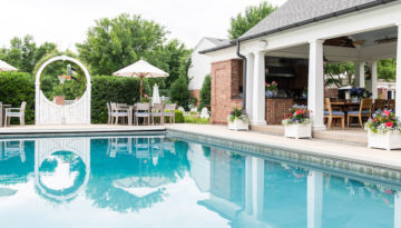 A wide, ocean-y blue pool is lined by a roofed patio with tables and chair and a full kitchen. Through shrubbery fences and a white gate with a decorative circle frame is a basketball hoop.