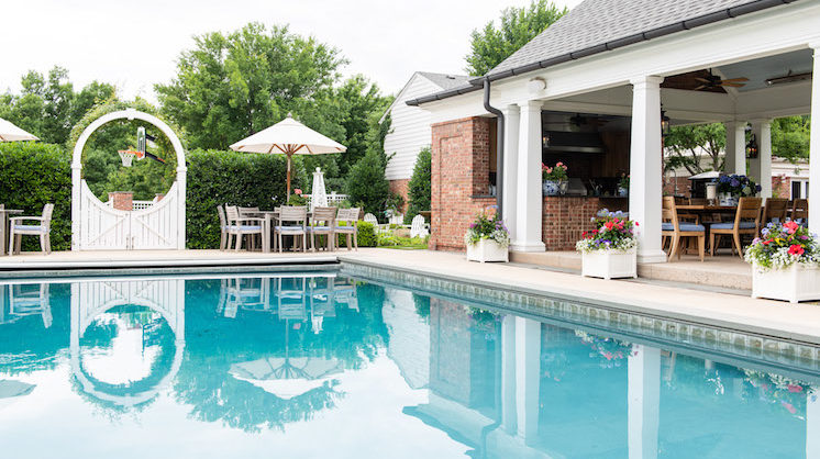 A wide, ocean-y blue pool is lined by a roofed patio with tables and chair and a full kitchen. Through shrubbery fences and a white gate with a decorative circle frame is a basketball hoop.