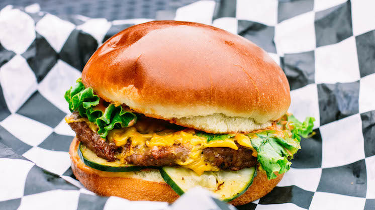 The Green Daze burger from Burger Punk on checkered paper. In between its shiny bun is a Beyond Meat patty, American cheese, house pickles and punk sauce.
