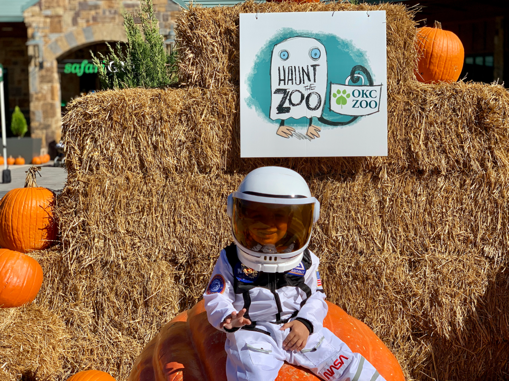 A small child in an astronaut costume sits on a massive pumpkin. Behind the child is a stack of hay with a sign attached reading, "Haunt the Zoo, OKC Zoo."
