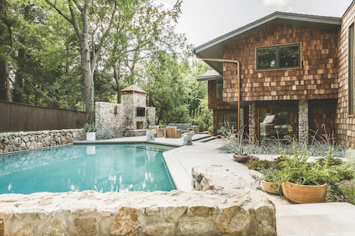 A blue pool is enclosed by stone walls. A wooden cabin with a similar stone foundation is behind the pool.