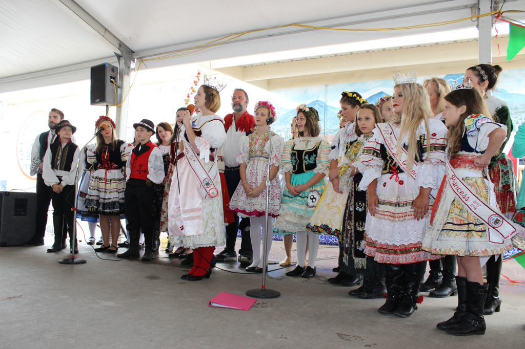 A group of children are dressed in traditional Czech clothing under an event tent. There are microphones and speakers set up.