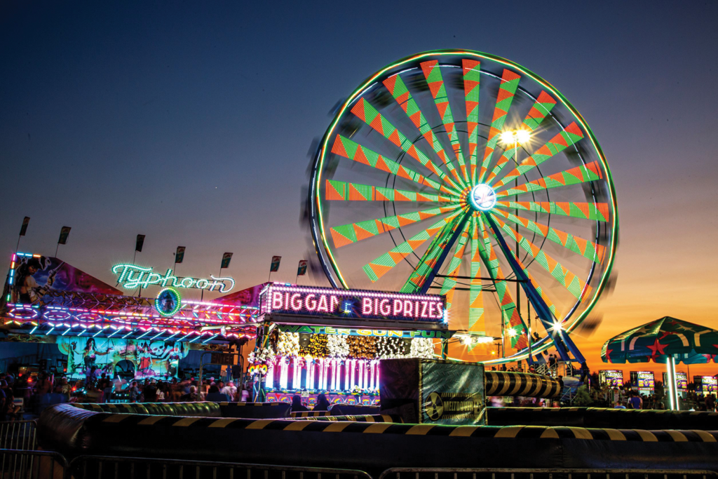 A green and orange Ferris wheel spins against a sky at dusk, with other fair booths and rides lit up in neon colors.