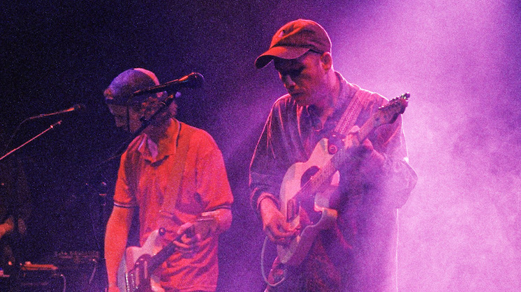 Wil Norton and Danny Davis of Husbands are on stage performing with guitars. Danny wears a red polo shirt, while Wil wears a baggy button-up. Both are wearing hats and gazing at their guitars pedals. A purple haze sifts through the stage.