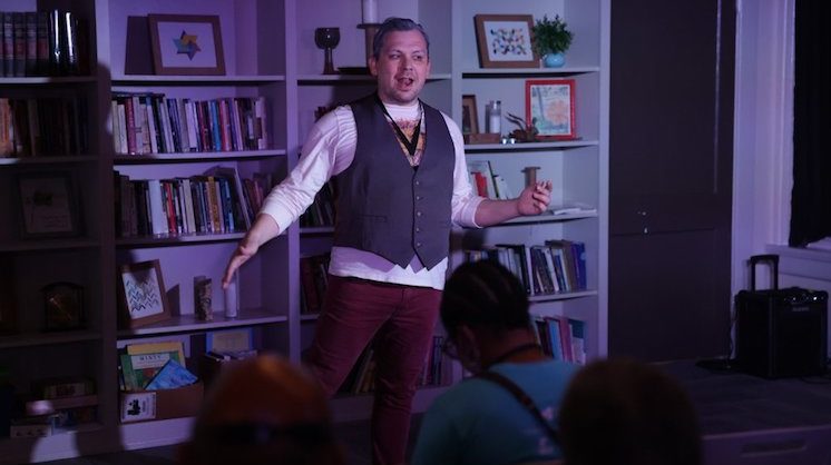 Actor and writer Rodney Brazil performs on a dark stage floor with a neat bookshelf behind him.