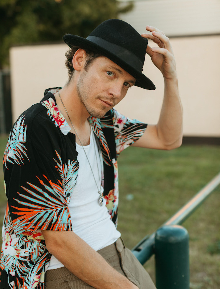 Rapper Josh Sallee, wearing a opened-up dress shirt with palm leaves, tips hit hat with his left arm while sitting on a bar outside.