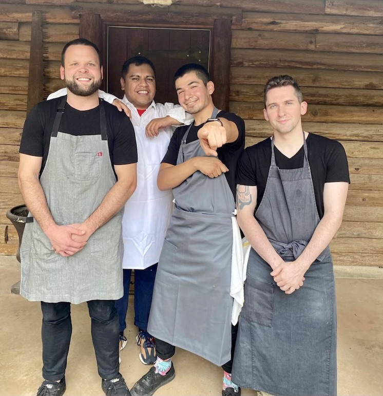 Chefs Joel Wingate, Roger Herrera, Jessie Gomez and Dylan Adams stand smiling and wearing aprons outside a wooden cabin.
