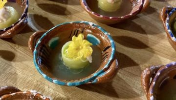 A citrus-y fruit adorned with a flower sits in a greenish liquid in an ornately decorated brown bowl with blue highlights.