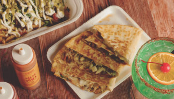 A plate of quesadillas from Trompudo's Tacos sits next to a green margarita and a styrofoam box of al pastor fries.