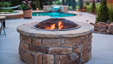 A circular stone fire pit holds little flames on a concrete outdoor patio. Shrubs, potted flowers and a cerulean blue waterfall and blue are behind the fire pit.