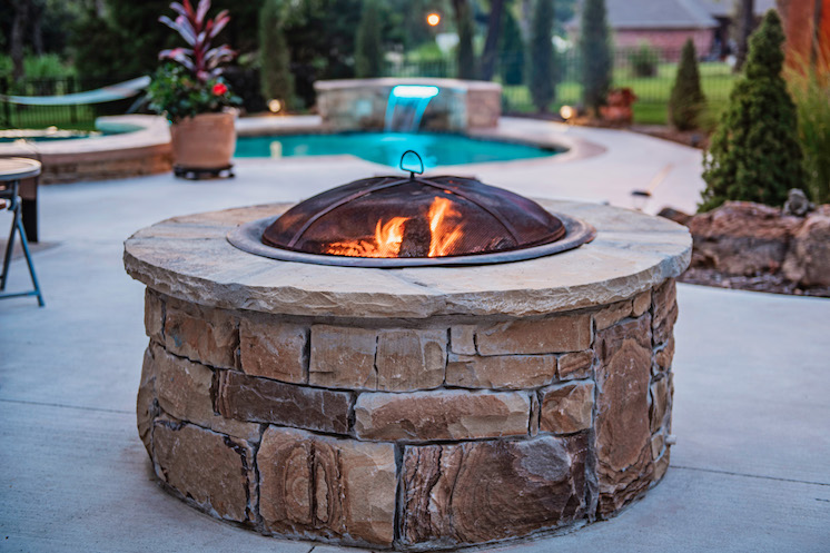 A circular stone fire pit holds little flames on a concrete outdoor patio. Shrubs, potted flowers and a cerulean blue waterfall and blue are behind the fire pit.