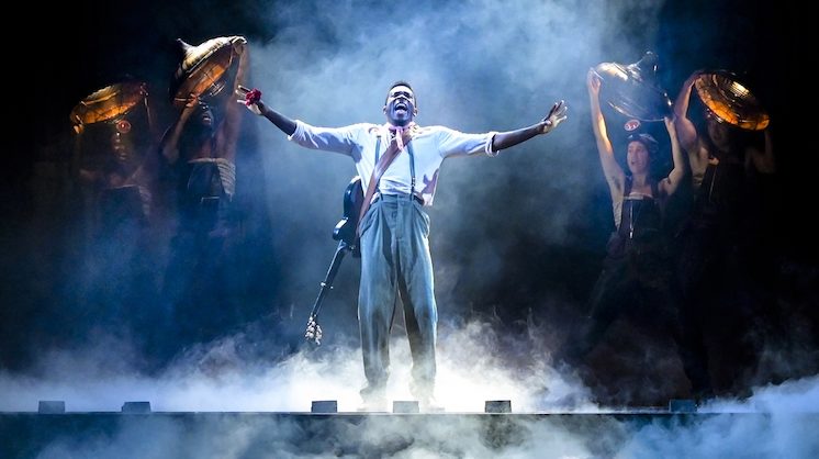 Chibueze Ihuoma exclaims on a foggy stage wearing a guitar on his back during a touring production of Hadestown.