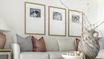 Three framed black and white photos hang above a white couch accented with brown and gray pillows on a white wall. A marbled ceramic pot holds thin branches of a plant.