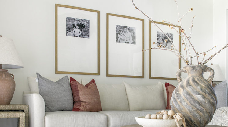 Three framed black and white photos hang above a white couch accented with brown and gray pillows on a white wall. A marbled ceramic pot holds thin branches of a plant.