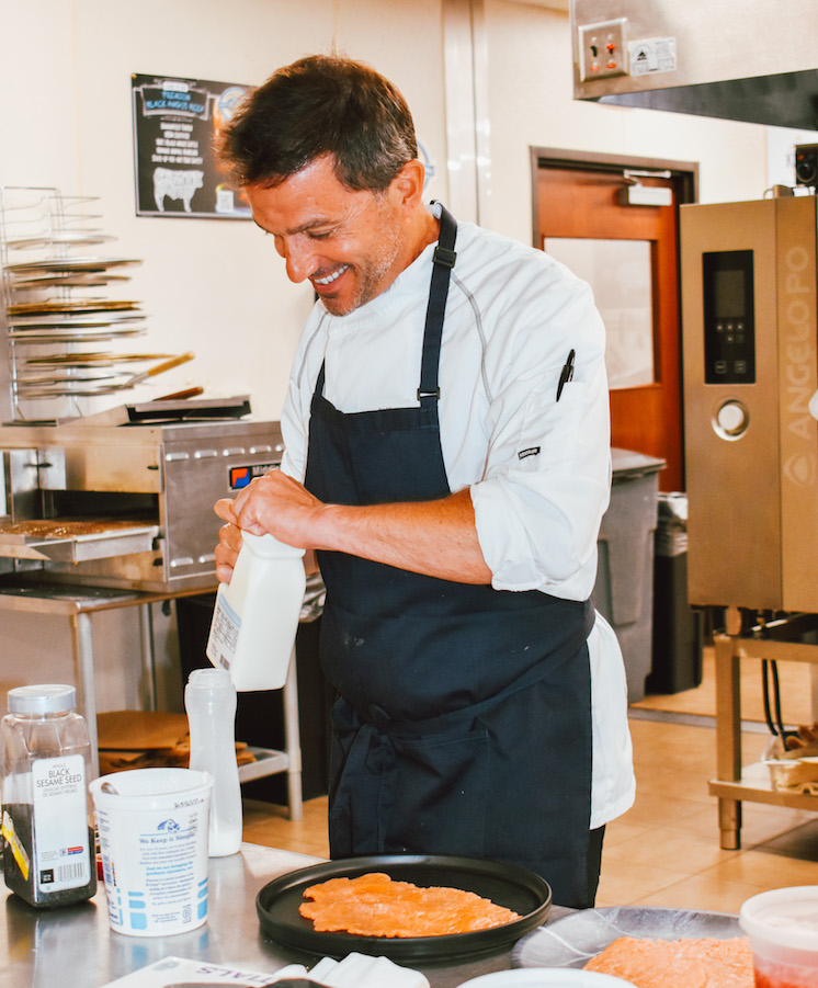 Executive Chef of NOUN Hotel Greg Lopez cooks in his kitchen, his head titled down and smiling.