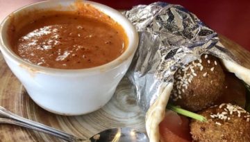 A gyro filled with falafel is wrapped in aluminum foil next to a red sauce.