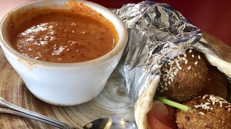 A gyro filled with falafel is wrapped in aluminum foil next to a red sauce.