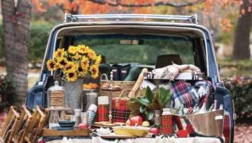 The bed of a truck is decorated with picnic baskets, plaid blankets, a vase of sunflowers, a tub of drinks and tailgating food. Orange autumnal trees hang in the background.