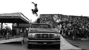 A skater wearing a ballcap, no shirt and black jeans skates over a Ford pickup truck in front of a mural at the Plaza District. Two wooden ramps lead up to the truck. A surrounding crowds marvels and takes videos of the trick.
