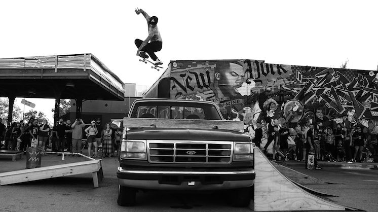 A skater wearing a ballcap, no shirt and black jeans skates over a Ford pickup truck in front of a mural at the Plaza District. Two wooden ramps lead up to the truck. A surrounding crowds marvels and takes videos of the trick.