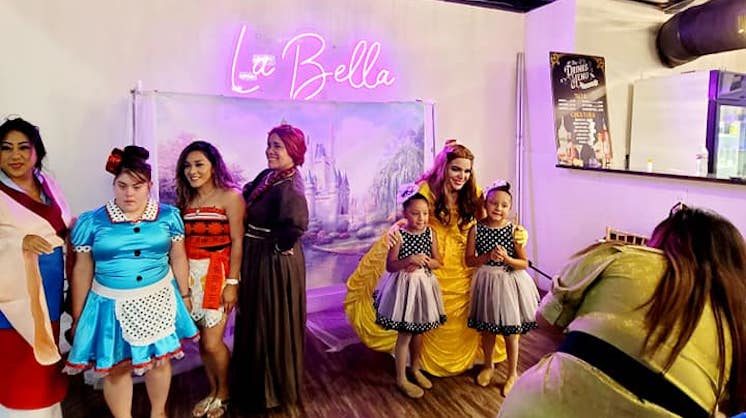 Young models pose for photos with experienced one in front of a backdrop illuminated with a neon sign reading "La Bella."