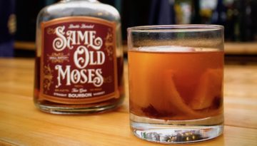 An old fashioned drink, complete with orange peel, sits on a wooden table next to a bottle of Same Old Moses whiskey.