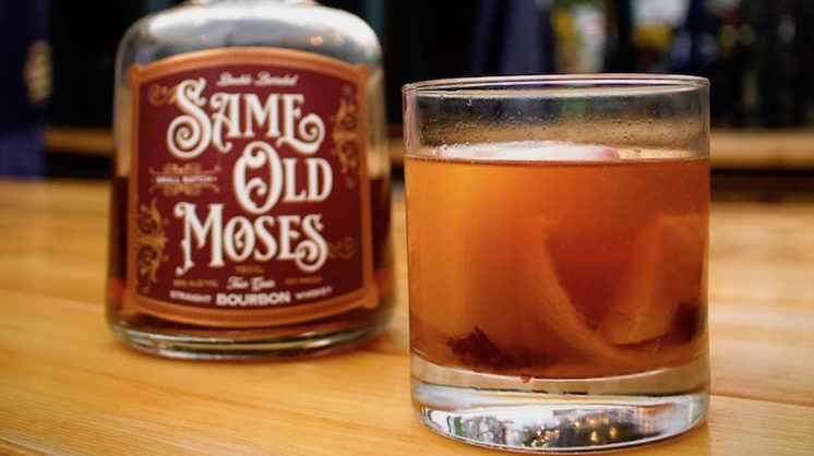 An old fashioned drink, complete with orange peel, sits on a wooden table next to a bottle of Same Old Moses whiskey.