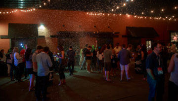 A scattered group of people stand outside at night with stringed bulb lights hanging above the events space.