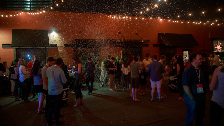 A scattered group of people stand outside at night with stringed bulb lights hanging above the events space.