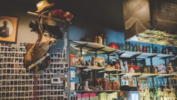 A mounted deer head on a bar wall wears sunglasses, a chain and an array of eclectic hats. Numerous Polaroids cover the wall. A rack of liquor at the bar is next to the pictures.