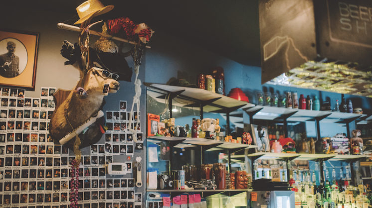 A mounted deer head on a bar wall wears sunglasses, a chain and an array of eclectic hats. Numerous Polaroids cover the wall. A rack of liquor at the bar is next to the pictures.