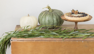 Two pumpkins, one green and one white, sit on wooden table next to an elevated platter with a fig tart and a fall grass hanging off the table.