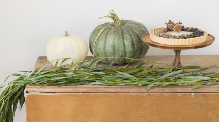 Two pumpkins, one green and one white, sit on wooden table next to an elevated platter with a fig tart and a fall grass hanging off the table.