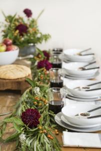 Clean white bowls are set in a line along a wooden table with wintry greens decorating the center of the table.