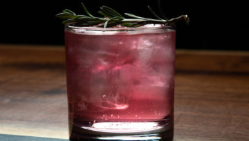A cloudy pink drink in a whiskey glass is topped with a green garnish. The glass sits on a wood tabletop.