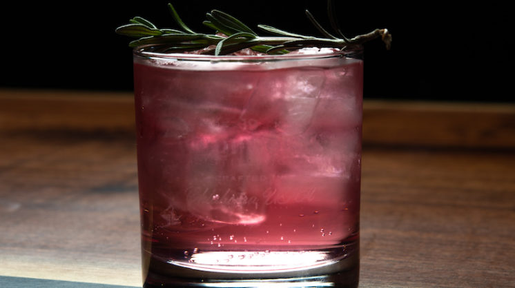 A cloudy pink drink in a whiskey glass is topped with a green garnish. The glass sits on a wood tabletop.