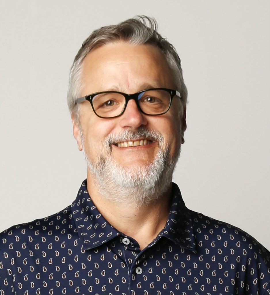 A smiling man in his 50s poses from the upper torso and up wearing a patterned navy button-up shirt with gray hair and beard and black glasses.