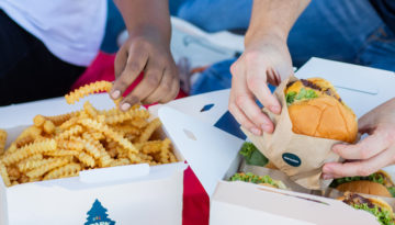 Two people dig into two takeout boxes. One box contains french fries and the other holds burgers.