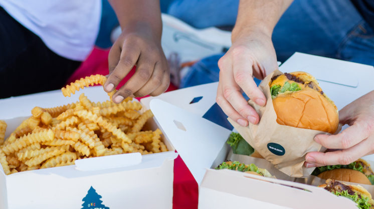 Two people dig into two takeout boxes. One box contains french fries and the other holds burgers.
