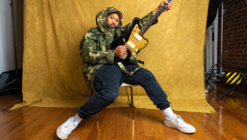 Indie rock musician Bartees Strange, wearing a camouflage hoodie, sits in a chair against a velvet yellow backdrop and strums his guitar in the air.