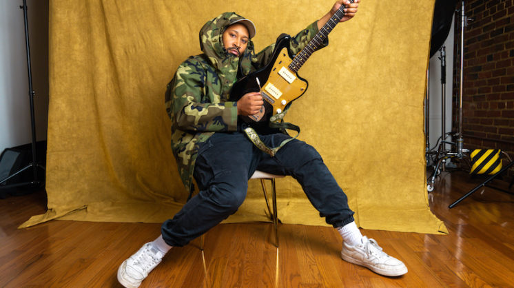 Indie rock musician Bartees Strange, wearing a camouflage hoodie, sits in a chair against a velvet yellow backdrop and strums his guitar in the air.