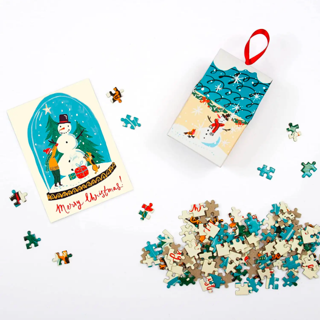 A box ornament that holds puzzle pieces that make up the image of a snow globe.