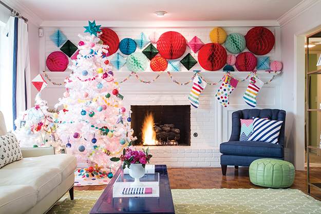 A white living room contains highlights of red, green, blue, orange and yellow circles, with a completely white Christmas tree decorated with ornaments of all colors and white stockings with rainbow stripes hanging on the fireplace.