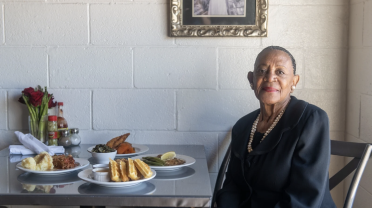 Award-winning chef Florence Jones sits next to her creations at a table against a white brick wall.