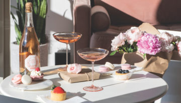 Two vintage glasses of sparkling rose sit on a table complete with baked sweets topped with strawberries and and pink flowers.
