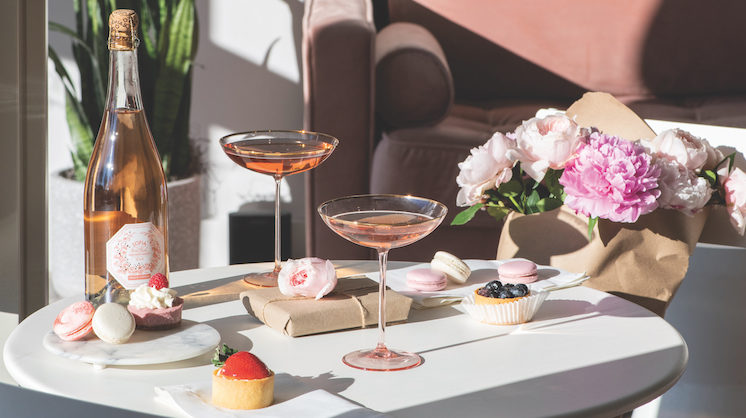Two vintage glasses of sparkling rose sit on a table complete with baked sweets topped with strawberries and and pink flowers.