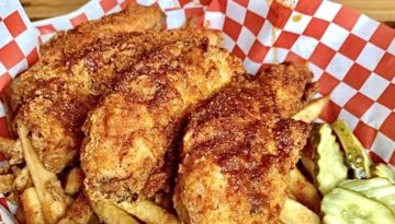 Three spiced chicken tenders sit next to french fries and sliced pickles in a checkered basket.