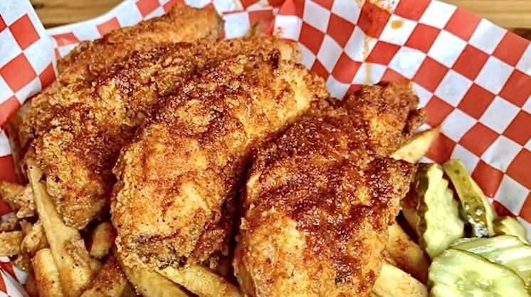 Three spiced chicken tenders sit next to french fries and sliced pickles in a checkered basket.
